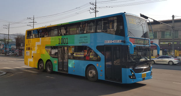 Ein blauer und gelber Doppeldeckerbus fährt auf einer Straße mit anderen Fahrzeugen, Strommasten, Gebäuden, Bäumen und einem klaren Himmel im Hintergrund.