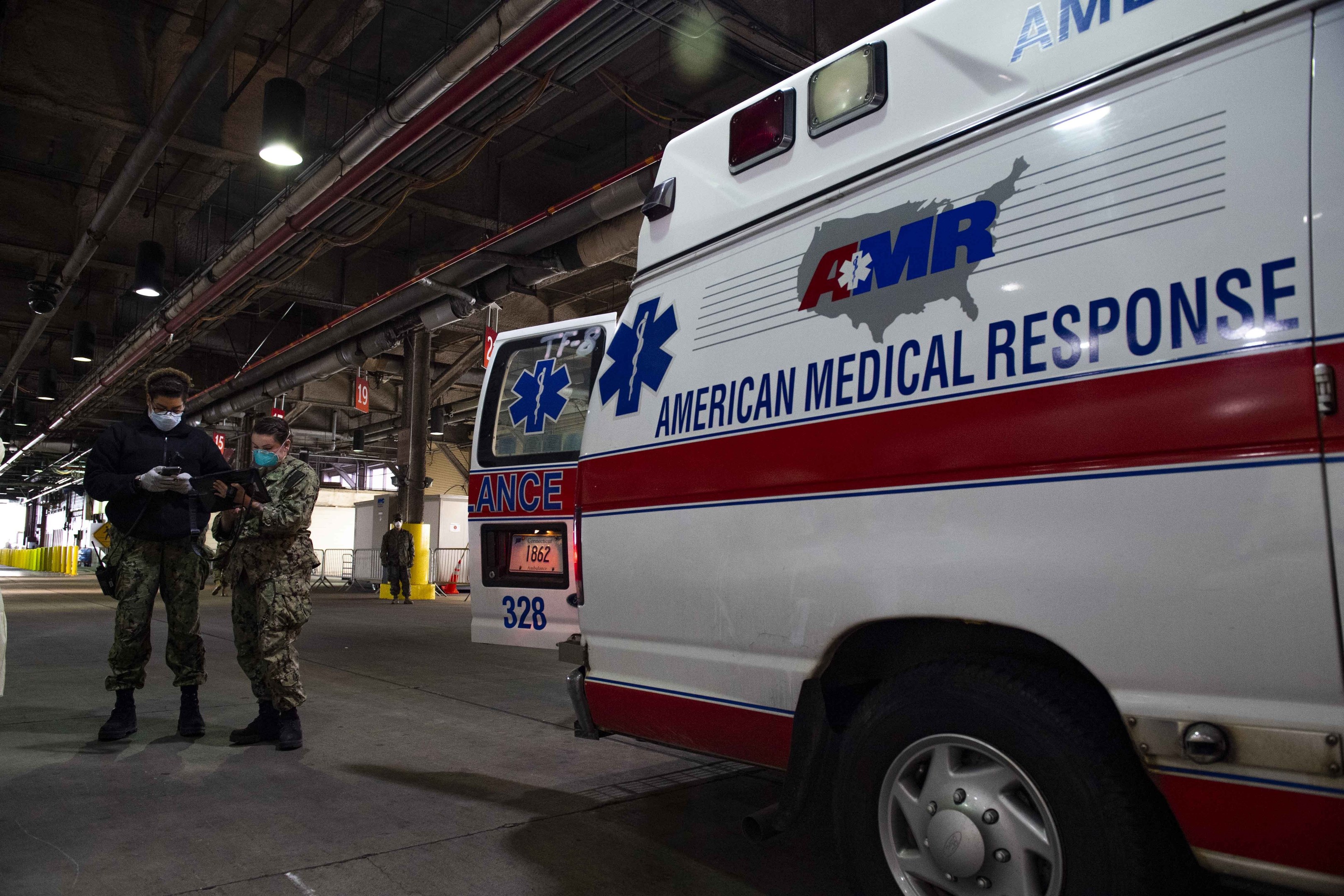American Medical Response ambulance parked indoors with two masked individuals in front of it, a vehicle to the left, and additional people and objects in the background.