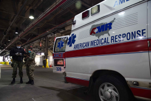 American Medical Response ambulance parked indoors with two masked individuals in front of it, a vehicle to the left, and additional people and objects in the background.