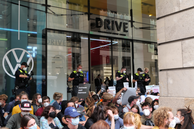 Eine große Gruppe von Menschen mit Masken und Schildern vor einem Glasgebäude, einige tragen Mützen und halten Kameras, was auf eine Demonstration in Melbourne, Australien, hindeutet.