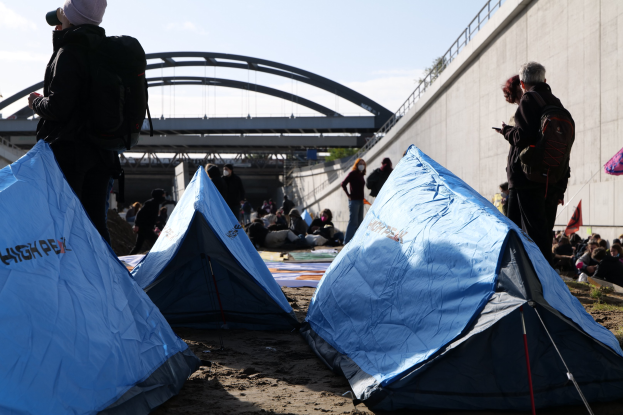 Eine Gruppe von Menschen sitzt auf einem sandigen Strand in der Nähe von Zelten, mit einer Wand und einer Brücke im Hintergrund, die an einer Klimawandel-Demonstration teilnehmen unter einem bewölkten Himmel.