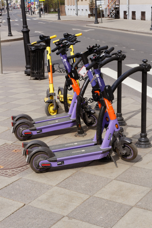 Eine Reihe von Elektro-Scootern, die auf einer städtischen Straße geparkt sind, mit einem Mülleimer im Vordergrund und Gebäuden, Bäumen und Fahrzeugen im Hintergrund.