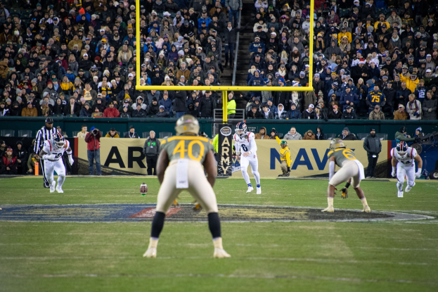 Ein Fussballspiel wird in einem Stadion mit Spielern in Sportkleidung und Helmen im Vordergrund und einer Zuschauermenge im Hintergrund sowie sichtbaren Pfosten und Schildern mit Text gespielt.