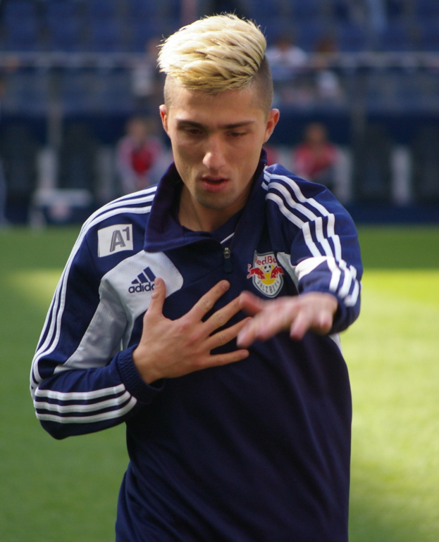 Ein junger Mann in einem blauen T-Shirt steht auf einem Fußballfeld mit ausgestreckten Armen und entschlossener Miene als Flügelspieler.