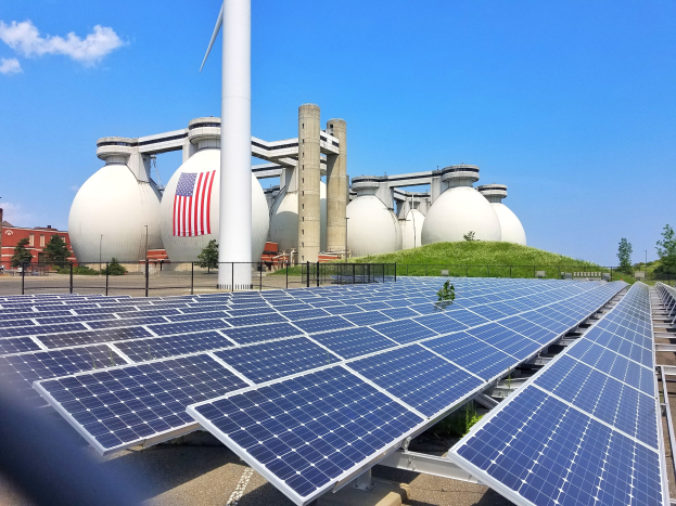 Solarpanels auf dem Dach eines Gebäudes mit einer Flagge, umgeben von einem Zaun, Gras, Pflanzen, Bäumen, Pfählen und anderen Gegenständen, mit zusätzlichen Gebäuden und einem bewölkten Himmel im Hintergrund.