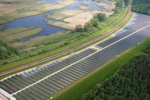 Luftaufnahme einer Solaranlage mit Solarpanelen auf einem Feld, umgeben von Bäumen, Gras, Pflanzen und Wasser, mit einem Zug auf einer nahen Eisenbahnstrecke.