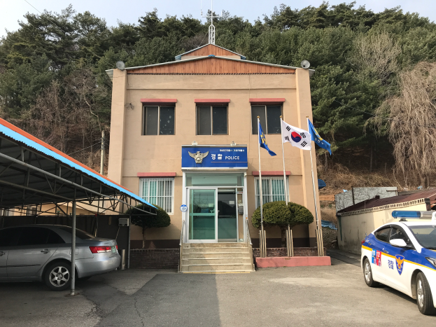 Ein Polizeiwagen vor dem Seoul Polizeipräsidium, ein mehrstöckiges Gebäude mit Fenstern, Türen, Stufen, Pflanzen, Fahnen auf Stangen, einem Schuppen und anderen Gegenständen, mit Bäumen und einem klaren blauen Himmel im Hintergrund.