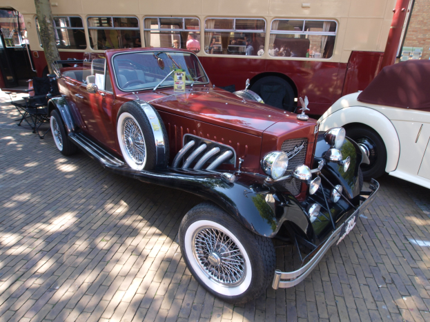 Ein Oldtimer neben einem Doppeldeckerbus auf einem Kopfsteinpflasterweg, mit Stühlen und einem Baum im Vordergrund, einem Gebäude mit Fenstern im Hintergrund und Menschen im Bus zu sehen.