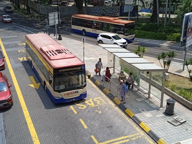 Stadtstraße mit fahrenden Autos, Bussen, einer Bushaltestelle, Fußgängern auf dem Gehweg, Strommasten, Schildern, Bäumen, Gras und Gebäuden im Hintergrund.