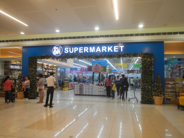 Eine Gruppe von Menschen vor einem Supermarkt, einige mit Einkaufstäschen, mit einem Einkaufswagen, Topfpflanze, Regalen, Schildern und Deckenleuchten im Hintergrund.