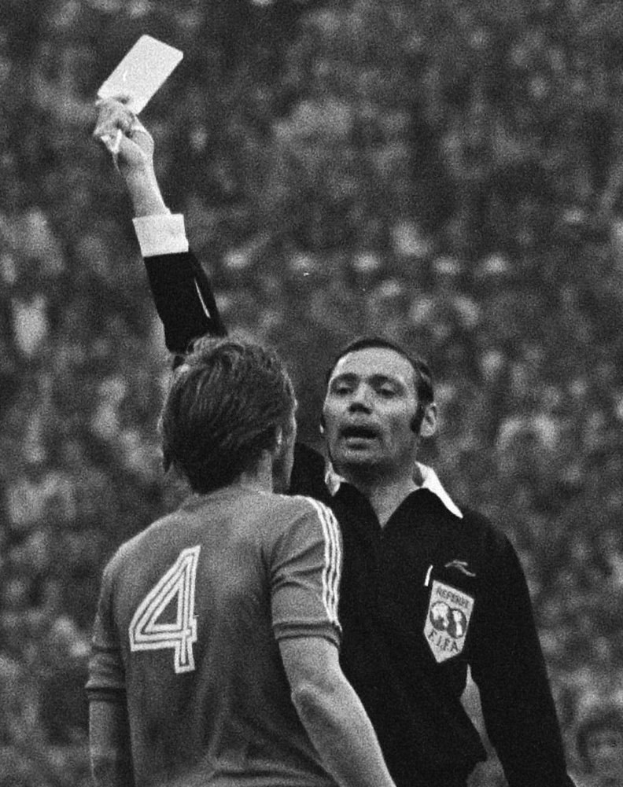 Ein Schiedsrichter in einem Schwarz-Weiß-Foto hält eine weiße Karte vor einem Fußballspieler, mit einem unscharfen Hintergrund.