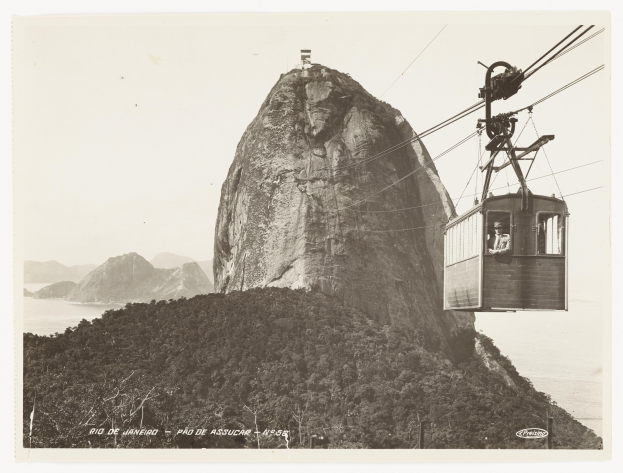 Schwarz-weiß-Foto einer Seilbahn, die einen Berg hinauffährt, mit einem Passagier darin, umgeben von Bäumen, Hügeln und Wasser; Text ist unten sichtbar.