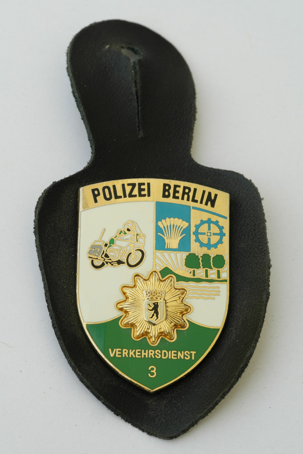 Ein schildförmiges Polizeibadgebnis mit "Polizei Berlin" in schwarzem Text auf einem blauen Hintergrund mit einem weißen Stern und Rahmen, das auf einer Oberfläche ruht.