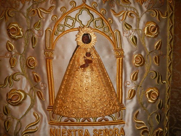 Goldbestickte Chasuble mit einer friedlichen Frau in einem langen Kleid mit hohem Kragen und Zopf, umgeben von detaillierten Designs, Text und goldenen Rändern.