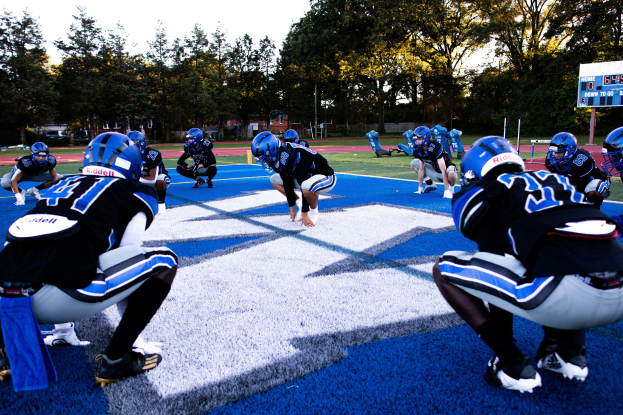 Junge Männer in Sportkleidung und Helmen beim Fußballspielen auf einem Feld mit einem Scoreboard und Bäumen im Hintergrund unter einem klaren blauen Himmel.