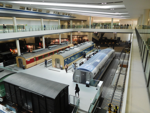 Eine großzügige Museumsausstellung zeigt verschiedene Züge in unterschiedlichen Größen und Farben, bei der Besucher von verschiedenen Ebenen und dem Erdgeschoss aus beobachten.