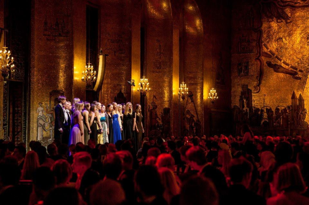 Eine Gruppe von Menschen auf einer beleuchteten Bühne mit bemalten Wänden im Royal Opera House, betrachtet von einem Publikum.