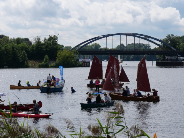 Eine Gruppe von Menschen in kleinen Booten auf einem Gewässer, mit einer Brücke, Fahrzeugen, Bäumen und einem bewölkten Himmel im Hintergrund und Pflanzen unten, die ein Regatta vermuten lassen.