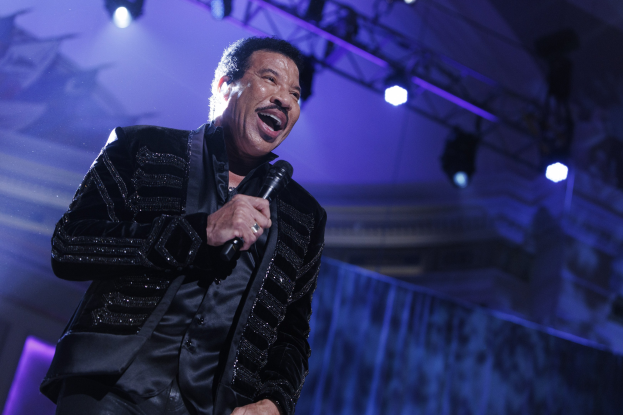 Lionel Richie performt auf den Grammys, trägt ein schwarzes Kleid und hält ein Mikrofon auf einer hell erleuchteten Bühne.