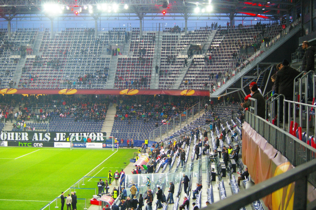 Großes Stadion voller Zuschauer bei einem Fußballspiel, mit sitzenden und stehenden Personen, einem Geländer, einer Fahne und einem leeren Hintergrund mit Deckenbeleuchtung.