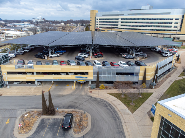 Luftaufnahme eines solarpanelbedeckten Parkplatzes mit geparkten Autos, umgeben von Gebäuden, Grünflächen, Straßen und Hügeln unter einem bewölkten Himmel.