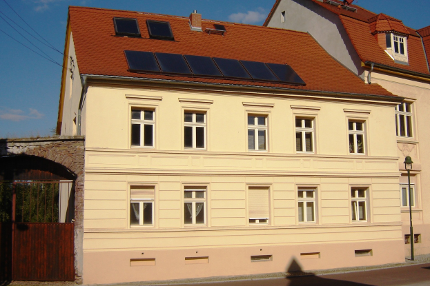 Gebäude mit Solar-Panelen auf dem Dach, umgeben von Straßeninfrastruktur einschließlich Masten, Laternen, Kabeln und Bäumen unter bewölktem Himmel.
