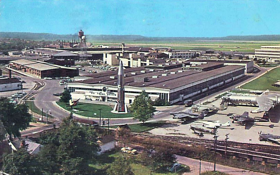 Altes Schwarz-Weiß-Foto eines Flughafens mit parkenden Flugzeugen, Gebäuden, Bäumen, Straßen, Fahrzeugen, Strommasten, Gras, Bahnschienen, Türmen, Hügeln und einem klaren Himmel, mit Text am unteren Rand.