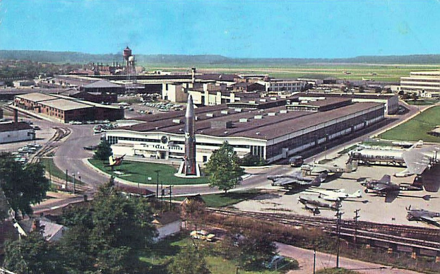 Altes Schwarz-Weiß-Foto eines Flughafens mit parkenden Flugzeugen, Gebäuden, Bäumen, Straßen, Fahrzeugen, Strommasten, Gras, Bahnschienen, Türmen, Hügeln und einem klaren Himmel, mit Text am unteren Rand.