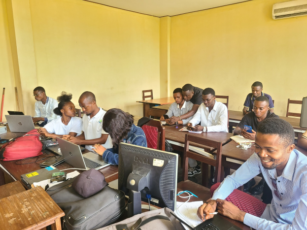 Gruppe von Menschen, die an Tischen in einem Klassenzimmer sitzen, mit Laptops, Taschen, Monitoren und Büchern auf den Tischen und einer Klimaanlage an der Wand im Hintergrund.