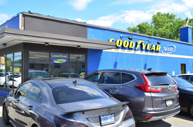 Außenansicht eines Goodyear Select Autohauses mit parkenden Fahrzeugen, Beschriftung auf dem Gebäude, Bäumen im Hintergrund und einer bewölkten Himmel.