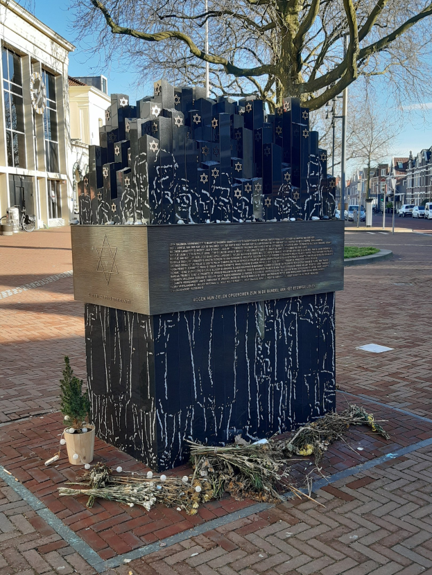 Denkmal in einem Stadtplatz gewidmet an Holocaust-Opfer, mit einem Baum davor, einer Topfpflanze auf dem Boden, verstreuten trockenen Blättern und Gebäuden, Fahrzeugen, Masten und Bäumen im Hintergrund.