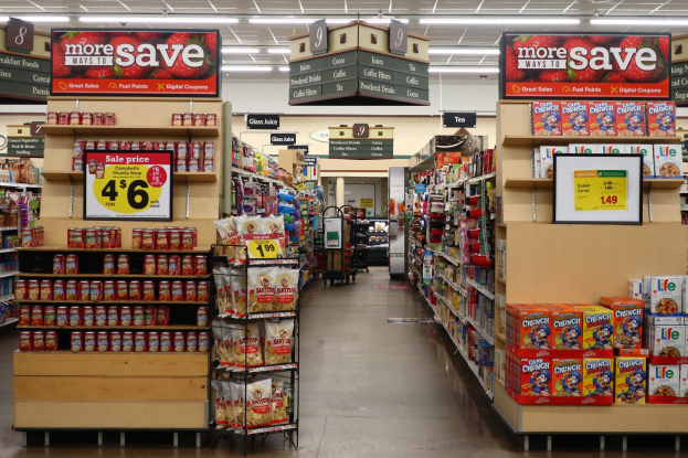Ein Kiosk-Innenraum mit Regalen und Boden voller verschiedener Lebensmittel, Deckenbeleuchtung und Informationsschilder im Hintergrund, mit einer Person in der Ferne sichtbar.
