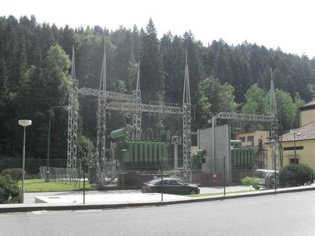 Eine elektrischen Umspannstation mit einem Zaun, Masten und Laternen, umgeben von Pflanzen, Gras und Bäumen, mit einem Auto auf der benachbarten Straße, Gebäuden im Hintergrund und einem klaren blauen Himmel.