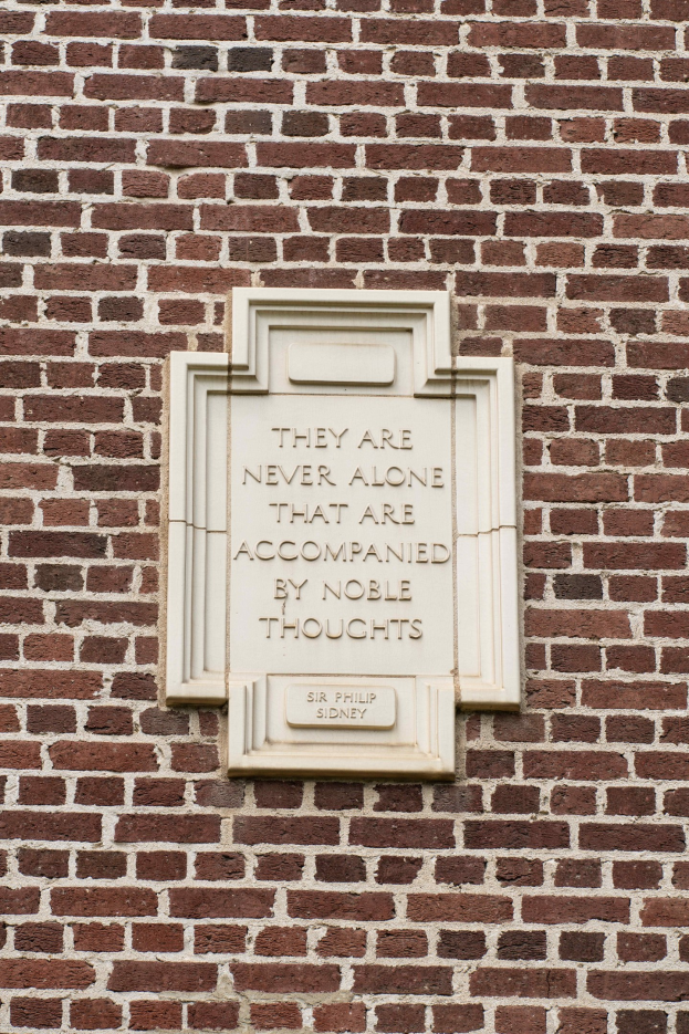 Eine Steinplatte auf einer Ziegelwand mit der eingravierten Inschrift "Sie sind nie allein, die von edlen Gedanken begleitet sind." in fetter Schrift.