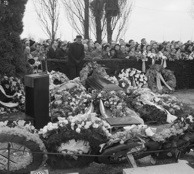 Schwarz-weißes Bild einer Trauerfeier mit einem Mann in einer Mütze, der auf einem Podium spricht, umgeben von Blumen, Stühlen und einer Menge, mit Bäumen und einem klaren Himmel im Hintergrund.