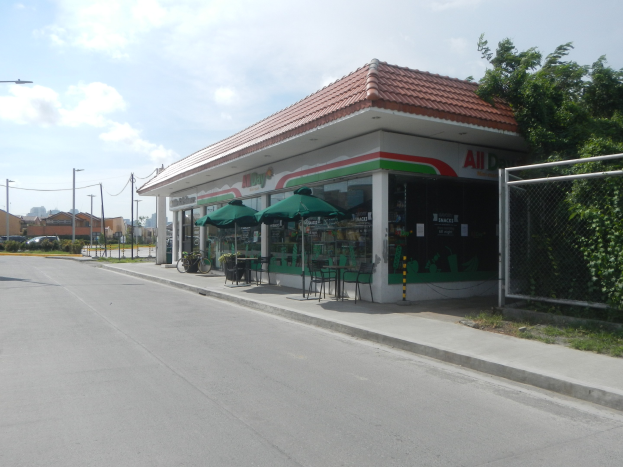 Laden an einer Stra├čenkreuzung mit einem Metallzaun, Vegetation, Fahrzeugen, n├Ąchsten Geb├Ąuden und einem bew├Âlktem Himmel; drinnen Tische mit Stühlen und Sonnenschirmen, einige mit Gegenst├Ąnden.