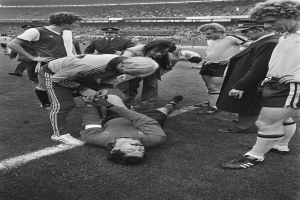 Schwarzes und weißes Bild eines Fußballspielers, der auf dem Boden liegt und von einer Gruppe Männer umgeben ist, mit einer Zuschauermenge im Hintergrund der Stadion.