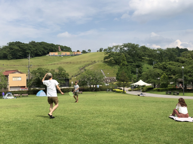 Eine Gruppe von Menschen, die Badminton in einem Park spielt, mit Zelten, Straßenlaternen, Strommasten, Stromkabeln, Gebäuden, Bäumen, Hügeln und einem bewölkten Himmel im Hintergrund; ein Mann hält einen Schläger in der Hand und sitzt auf einem Tuch neben einer Frau.