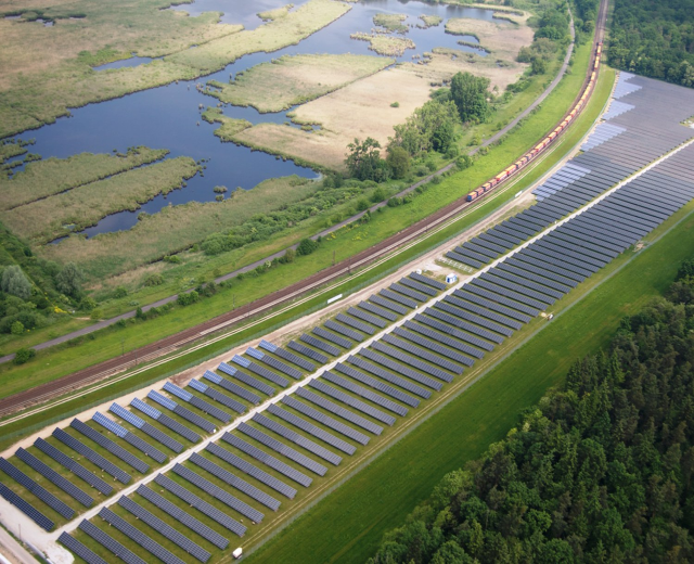 Luftaufnahme eines Solarparks mit Panelen in einem Feld, umgeben von Bäumen, Gras, Pflanzen und Wasser, mit einem Zug auf einer nahen Bahnstrecke.