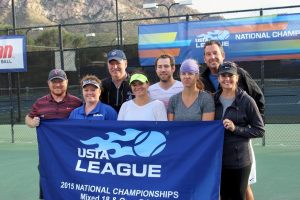 Gruppe von Menschen auf einem Tennisplatz mit einem "2015 USTA National Championships"-Schild, einem Zaun, Bannern, Laternenpfählen, Bäumen, Bergen und einem klaren blauen Himmel im Hintergrund.