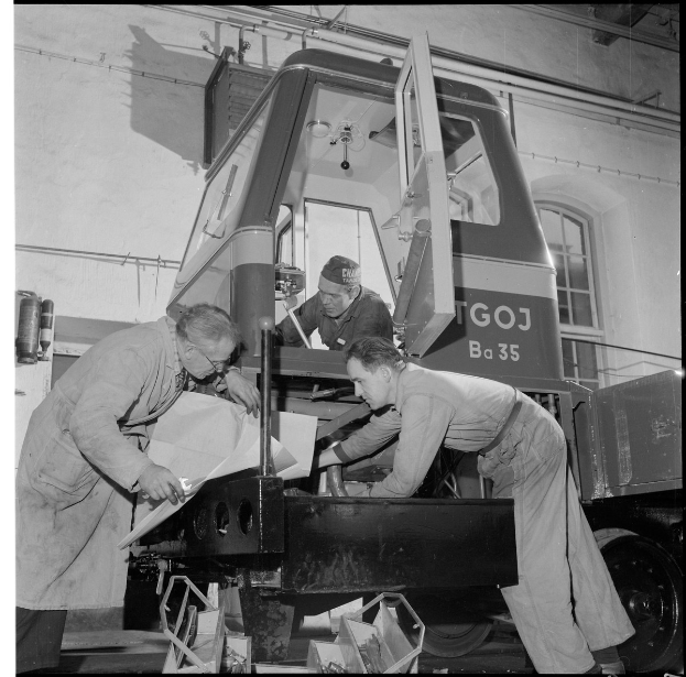 Drei Männer arbeiten an einem Lastwagen in einer Schwarz-Weiß-Fabrikumgebung mit verstreuten Gegenständen und einem Fenster im Hintergrund.
